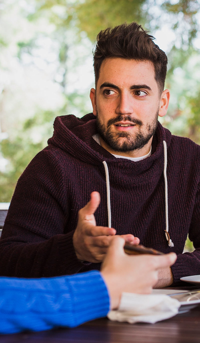 Man in a dark hoodie sitting at a table with another person, talking about the best way to quit smoking