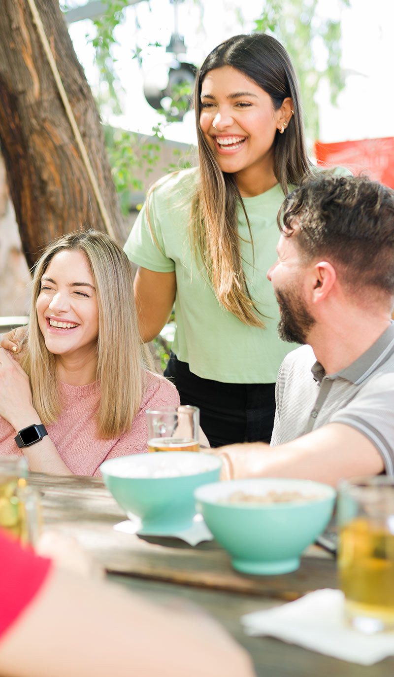 Two women and a man sitting at a table with drinks and food, smiling outdoors.