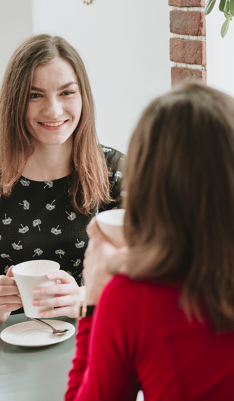 Two women sitting at a table, one holding a cup, in a casual setting.