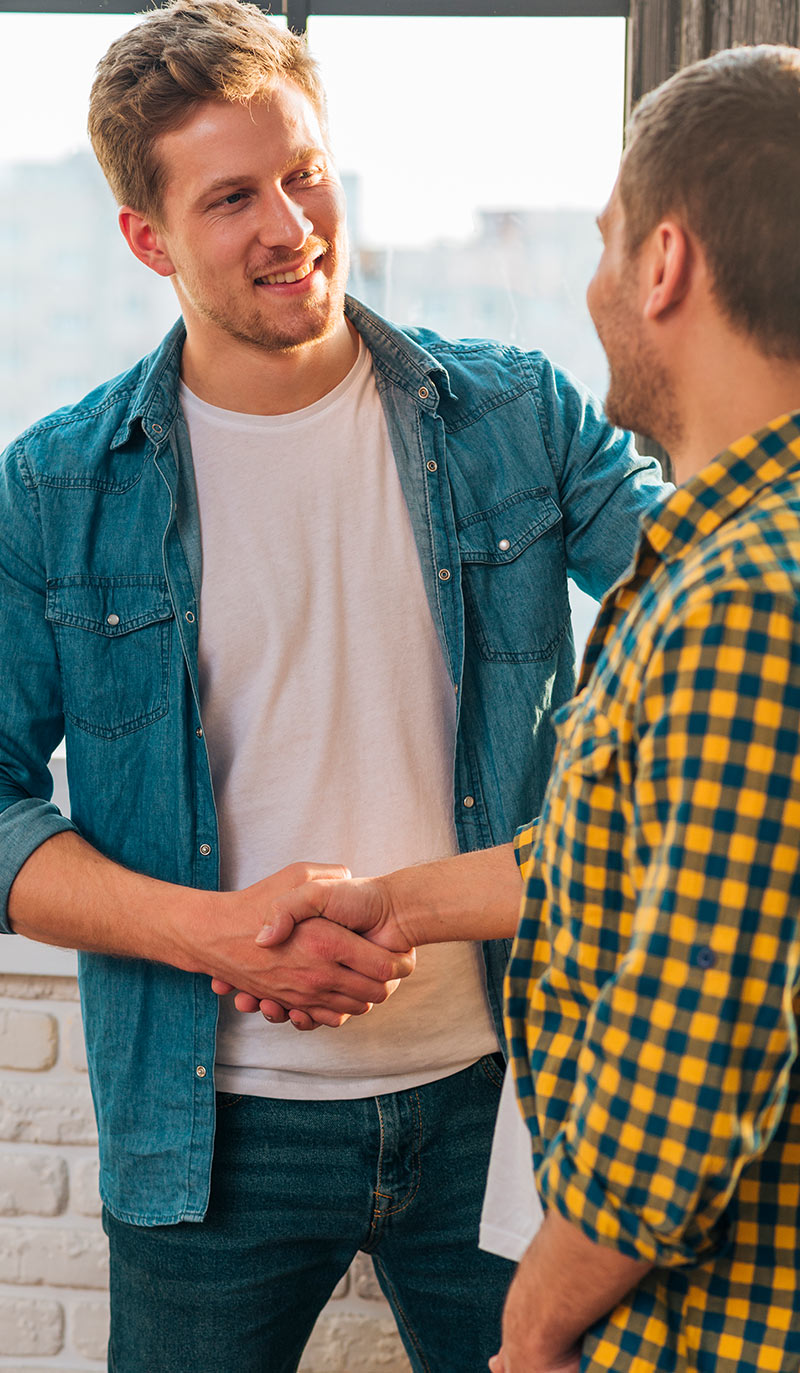 Two men shaking hands in a casual setting with a window and light-colored wall in the background.